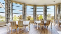 Located at 7600 S Mason Rd in Houston, Texas, this well-appointed dining room offers a refined and inviting atmosphere designed for both comfort and functionality. The space is defined by its striking floor-to-ceiling windows arranged in a graceful curved formation, flooding the interior with an abundance of natural light and providing expansive views of the surrounding greenery and outdoor landscape. Elegant floral-patterned drapes frame the windows, adding a touch of classic sophistication to the bright, airy environment.

The room features warm, wood-look flooring that runs throughout, complementing the neutral beige tones of the walls and creating a cohesive, welcoming aesthetic. Multiple white dining tables with ornately crafted legs are arranged throughout the space, accompanied by upholstered wooden chairs in a French country style — ideal for group gatherings or collaborative use.

Soaring ceilings with a ceiling fan enhance the sense of openness and volume, while wall-mounted sconces and a flat-screen television contribute to the room's versatility. A built-in cabinetry station with a countertop provides practical utility along one wall. Dark-framed interior doors with clean white trim add architectural contrast, and large mirrored panels further amplify the sense of space and light throughout this thoughtfully designed dining area.