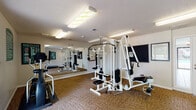 Located at 6900 Arbors Ct in Evansville, Indiana, this fitness center offers a well-equipped, single-floor exercise facility designed to support a range of workout routines. The space features carpet tile flooring throughout and is illuminated by overhead fluorescent lighting, creating a bright and functional environment for physical activity.

The room is anchored by a multi-station cable weight machine with multiple cable pulleys, weight stacks, and adjustable seats, allowing users to target a variety of muscle groups. Cardio equipment includes multiple treadmills — including a Pro Maxima model — and an elliptical trainer, providing comprehensive options for aerobic exercise. A wall-mounted coat hook rail offers convenient storage for personal items.

A large floor-to-ceiling mirror spans one wall, visually expanding the space and allowing users to monitor their form during workouts. Motivational fitness posters and instructional charts are displayed on the walls, adding an informative and encouraging atmosphere to the room.

Natural light and outdoor views are accessible through a set of glass double doors, which open to an exterior walkway and green landscaping, enhancing the overall feel of the space. A fitness center rules and guidelines sign is posted near the entrance. The textured ceiling and clean white walls complete the practical, community-oriented design of this well-maintained fitness facility.