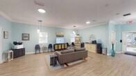 This single-floor space offers a generously proportioned, open-plan layout finished with light hardwood-style flooring and soft aqua-blue walls, creating a calm and welcoming atmosphere. Crown molding runs along the perimeter of the white ceiling, lending an elevated, polished character to the interior. Illumination is provided by a combination of recessed ceiling lights, modern pendant fixtures, and a starburst chandelier, ensuring the space is bright and well-lit throughout.

The main area is organized into several distinct functional zones. A central lounge area features a contemporary gray sofa set on a layered area rug, oriented toward a flat-screen television mounted atop a cane-front media console. Nearby, a cluster of sleek black chairs and small bistro tables provides additional seating. A cozy conversational nook is furnished with tufted yellow-green accent chairs arranged around a round table, accented by framed coastal artwork on the walls.

Along one wall, a beverage and refreshment station includes a credenza, water cooler, small beverage refrigerator, and coffee maker. A dedicated workstation with a desk and ergonomic chair serves administrative functions. Multiple windows allow natural light to filter in, enhancing the airy, open feel of the space.