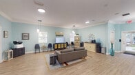 This single-floor space offers a generously proportioned, open-plan layout finished with light hardwood-style flooring and soft aqua-blue walls, creating a calm and welcoming atmosphere. Crown molding runs along the perimeter of the white ceiling, lending an elevated, polished character to the interior. Illumination is provided by a combination of recessed ceiling lights, modern pendant fixtures, and a starburst chandelier, ensuring the space is bright and well-lit throughout.

The main area is organized into several distinct functional zones. A central lounge area features a contemporary gray sofa set on a layered area rug, oriented toward a flat-screen television mounted atop a cane-front media console. Nearby, a cluster of sleek black chairs and small bistro tables provides additional seating. A cozy conversational nook is furnished with tufted yellow-green accent chairs arranged around a round table, accented by framed coastal artwork on the walls.

Along one wall, a beverage and refreshment station includes a credenza, water cooler, small beverage refrigerator, and coffee maker. A dedicated workstation with a desk and ergonomic chair serves administrative functions. Multiple windows allow natural light to filter in, enhancing the airy, open feel of the space.