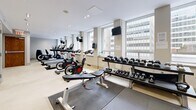 This fitness center is a comprehensively outfitted facility designed to support a full range of exercise routines. The space features a clean, modern aesthetic with neutral-toned walls, recessed and track lighting, and light-colored flooring that creates a bright, open atmosphere throughout.

The cardio zone is well-stocked with multiple Life Fitness treadmills, stationary cycling bikes, and additional cardio equipment, all positioned to take advantage of large windows that allow natural light to filter in and offer views of the surrounding urban environment. Mounted televisions provide entertainment during workouts.

The strength training area includes a multi-functional cable machine for a variety of resistance exercises, along with a full rack of dumbbells spanning a wide range of weights. Rubber floor mats are laid beneath the free weight stations for safety and durability. Stability balls in multiple sizes are neatly stored along one mirrored wall, which also serves to visually expand the space.

Full-length mirrors line a dedicated section of the facility, ideal for form-checking during workouts. A water cooler station is conveniently accessible, and a wall-mounted defibrillator ensures safety standards are met. The space also includes a separate access door for easy entry and exit. Overall, the fitness center provides a practical, well-maintained environment suited for diverse training needs.