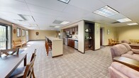 Located at 6940 Woodmere Rd in Woodbury, MN, this single-floor community space offers a generous and versatile layout designed to accommodate a wide range of social and recreational activities. The open floor plan flows seamlessly across distinct functional zones, all finished with neutral carpeting and drop-tile ceilings fitted with bright panel lighting throughout.

The game room area serves as a central highlight, featuring a full-size billiards table and a ping-pong table, providing ample space for play without crowding. A lounge area offers comfortable seating centered around a striking stacked-stone fireplace with a wall-mounted television above, creating a warm and inviting gathering spot. Natural light filters in through multiple windows, offering views of the surrounding greenery.

The kitchen is well-equipped with dark wood cabinetry, stainless steel appliances including a refrigerator, stove, and dishwasher, and an extended island with bar-height seating — ideal for catering or casual dining. An adjacent dining area with tables and chairs comfortably accommodates groups. A hallway connects the various sections of the space, which also includes a smaller seating nook with hardwood-style flooring, adding a cozy retreat from the main activity areas. This space is well-suited for community events, social gatherings, and recreational use.