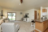 This single-floor residence blends practical design with charming farmhouse-style touches throughout. The living area is carpeted and comfortably sized, featuring a sliding glass door that fills the space with natural light and provides a view of the surrounding greenery. A wall-mounted air conditioning unit and baseboard heating ensure year-round comfort. The room opens naturally toward the kitchen, creating a functional flow between living and cooking spaces.

The galley-style kitchen is efficiently laid out with warm oak cabinetry, dark laminate countertops on both sides, and hardwood-style flooring. It includes a double basin sink, white appliances including a range and dishwasher, and a closet pantry at the end of the corridor, offering ample storage.

The primary bedroom is generously proportioned, featuring carpet flooring, a ceiling fan with light fixture, a window with outdoor views, and baseboard heating. The second bedroom also offers carpeted flooring and a ceiling fan. Both rooms carry a cohesive farmhouse aesthetic with distressed white wood furniture accents.

Two bathrooms serve the unit. The full bath includes a tub and shower combination with sliding glass doors and tile flooring. The second bathroom features a single-sink vanity with a wall mirror and wood cabinetry. Both are clean and functional in design.