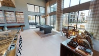 Located along FM-544 in Lewisville, Texas, this single-floor community clubhouse is designed with a modern aesthetic and a refined mix of materials and textures. The game lounge commands attention with soaring double-height ceilings, floor-to-ceiling black-framed windows, and abundant natural light that floods the polished white tile floors. A sleek billiard table anchors the space beneath a dramatic cluster pendant light fixture, while woven textile wall art and glass doors opening to an outdoor patio contribute to the space's welcoming character. Adjacent to the game area, a comfortable lounge features oversized windows with plaid drapery panels, offering views of the surrounding community. An eclectic mix of furnishings — including a rustic pallet-style coffee table, patterned area rug, and media console — creates a relaxed yet curated atmosphere. The wet bar and gathering area showcase striking design details, including a richly veined marble island with bar seating, a wood coffered ceiling, geometric tile-accent walls in teal and terra cotta, and elegant pendant lighting. Built-in cabinetry and a coffee station complete this social hub, making it an ideal space for both casual interaction and community events.
