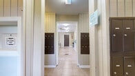 This single-floor community facility is efficiently organized into three functional areas connected by a continuous corridor with light beige tile flooring throughout. The entry section serves as a mailroom, flanked on both sides by rows of dark-finished individual lockboxes mounted on vertical-paneled walls, providing secure mail access for residents. Overhead fluorescent lighting ensures clear visibility in this area.

Beyond the mailroom corridor, the space opens into a well-equipped laundry room, which forms the centerpiece of the facility. Multiple top-loading washing machines and dryers are neatly arranged along the walls, each fitted with digital payment terminals for convenient operation. Natural light filters in through several windows offering views of surrounding greenery, complemented by ceiling-mounted fluorescent fixtures. A large folding table provides ample workspace for laundry tasks.

The third area functions as a utility and transitional space, featuring additional storage lockers, a folding table, and a dark-paneled exit door marked with safety signage including a fire extinguisher and exit indicator. The walls throughout are finished in warm beige tones with white wainscoting details, lending a clean, practical ambiance consistent across all areas of the facility.