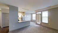 This single-floor space showcases an open-concept design that seamlessly connects the kitchen and living area. The kitchen is partially enclosed by a half-wall peninsula, which doubles as a breakfast bar with a built-in sink and offers a natural division between the two spaces. A track lighting system and elegant pendant lights hang above the peninsula, providing focused task lighting. The kitchen is equipped with stainless steel appliances, including a refrigerator and range, and features white cabinetry with decorative upper cabinet detailing. Recessed lighting in the kitchen adds additional illumination. The living area is finished with wall-to-wall carpeting, creating a warm and comfortable atmosphere. Neutral gray walls throughout lend the space a clean, modern aesthetic. Large windows allow ample natural light to fill the room, complemented by a glass-panel exterior door that further enhances brightness and provides direct access to the outdoors. The open layout and consistent neutral palette create a cohesive, airy feel throughout the space.