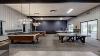 Located at 1400 W Third St in Chico, California, this single-floor community living room is a generously sized recreational space designed to foster social interaction and leisure. The room's industrial-modern aesthetic is defined by a mix of grid-patterned tile walls in neutral tones, bold red accent walls, exposed dark ceiling beams, and a combination of carpeted and hard-surface flooring that zones the space effectively.

The layout accommodates a wide variety of activities. Multiple pool tables take center stage beneath stylish geometric pendant light fixtures, while a ping-pong table provides additional competitive entertainment. A dedicated dartboard station with bar-height seating offers a classic game room experience. Vintage arcade cabinets line one wall, adding a retro gaming element to the space.

Comfortable seating areas — featuring upholstered sofas, lounge chairs, and low coffee tables — provide relaxed gathering spots throughout. Wall-mounted flat-screen televisions enhance the entertainment options. Potted plants are distributed throughout, softening the industrial finishes and adding a touch of natural warmth. Large sliding glass doors and windows allow ample natural light to fill the space and provide views of the surrounding outdoor area, contributing to an open, inviting atmosphere throughout.