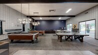 Located at 1400 W Third St in Chico, California, this single-floor community living room is a generously sized recreational space designed to foster social interaction and leisure. The room's industrial-modern aesthetic is defined by a mix of grid-patterned tile walls in neutral tones, bold red accent walls, exposed dark ceiling beams, and a combination of carpeted and hard-surface flooring that zones the space effectively.

The layout accommodates a wide variety of activities. Multiple pool tables take center stage beneath stylish geometric pendant light fixtures, while a ping-pong table provides additional competitive entertainment. A dedicated dartboard station with bar-height seating offers a classic game room experience. Vintage arcade cabinets line one wall, adding a retro gaming element to the space.

Comfortable seating areas — featuring upholstered sofas, lounge chairs, and low coffee tables — provide relaxed gathering spots throughout. Wall-mounted flat-screen televisions enhance the entertainment options. Potted plants are distributed throughout, softening the industrial finishes and adding a touch of natural warmth. Large sliding glass doors and windows allow ample natural light to fill the space and provide views of the surrounding outdoor area, contributing to an open, inviting atmosphere throughout.