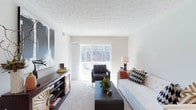 Located at 5146 N High St in the Clintonville neighborhood of Columbus, Ohio, this single-floor residence offers a well-designed layout filled with natural light and thoughtful details throughout. The living area serves as the centerpiece of the space, featuring plush carpeting, a textured ceiling, and a large sliding glass door that floods the room with daylight and provides direct access to a private balcony overlooking a lush, tree-lined backdrop. The generous proportions of the living room comfortably accommodate a full seating arrangement, making it an inviting space for both relaxing and entertaining. The neutral color palette of white walls and light carpeting creates a bright, airy atmosphere that allows for versatile interior styling. Bold decorative artwork and modern furnishings complement the clean architectural lines of the space. The balcony extends the living area outdoors, offering a serene view of mature trees that provide a natural sense of privacy and tranquility. The single-floor layout ensures convenient, step-free living throughout the residence. Overall, this space combines functional design with a comfortable ambiance, situated in one of Columbus's most established and walkable neighborhoods.