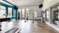 Located at 2951 Satellite Blvd in Duluth, GA, this fitness center offers a well-appointed, single-floor exercise space designed to support a full range of workout needs. The room features warm wood-look flooring throughout, crown molding, and a bold teal accent color scheme that energizes the space. Large multi-pane windows allow abundant natural light to flow in, complementing the recessed and track lighting overhead. Ceiling fans help maintain a comfortable environment during workouts.

The cardio zone is equipped with multiple treadmills, elliptical machines, and additional aerobic equipment, all positioned to take advantage of the natural light and garden views outside. A strength training area houses a cable machine, a leg press, a leg extension machine, and a power rack with a bench, catering to a variety of resistance training routines. A dedicated free weights section includes a tiered dumbbell rack with a broad range of weights, medicine balls, and TRX suspension straps anchored to the wall. Floor-to-ceiling mirrors line one wall, enhancing the sense of space and aiding in form correction. Wall-mounted TVs provide entertainment throughout the workout area, and built-in shelving offers convenient storage for personal items.