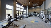 The fitness center at 100 Danforth Ave in Dobbs Ferry, New York, is a thoughtfully designed, single-floor facility that combines modern aesthetics with comprehensive workout amenities. The space is defined by soaring ceilings clad in warm wood paneling, which contrast beautifully with the cool gray tones of the surrounding walls, creating a refined yet energizing atmosphere. A generous row of floor-to-ceiling windows along one wall floods the interior with natural light and offers views of the surrounding tree-lined landscape, enhancing the overall workout experience. The open layout accommodates a wide variety of fitness equipment, including treadmills, ellipticals, stationary bikes, rowing machines, and an extensive selection of strength-training machines and free weights. Full-length mirrors line one side of the space, maximizing the sense of openness and allowing members to monitor their form during exercise. Overhead, large industrial-style ceiling fans and track lighting ensure comfortable air circulation and optimal visibility throughout. The flooring features a patterned carpet tile design with accents of yellow and blue, adding a touch of color and defining workout zones. A wall-mounted television is also available, offering entertainment during training sessions. This well-equipped fitness center delivers a motivating environment for residents and members pursuing their health and wellness goals.