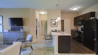 This single-story residence offers a thoughtfully designed open-concept floor plan that promotes a natural flow between the living, dining, and kitchen areas. The kitchen serves as a central highlight, featuring a spacious island topped with light granite countertops, dark espresso cabinetry, and pendant lighting that adds a refined touch overhead. Bar-height seating along the island creates an inviting space for casual dining or entertaining. The kitchen transitions seamlessly into the living area, which is finished with plush carpeting and receives warm, ambient light throughout. Walls in a soft neutral tone contribute to the bright and welcoming atmosphere of the space. A hallway visible from the main living area leads to additional rooms, hinting at a well-organized and functional layout beyond the open living space. Built-in shelving provides convenient storage, while framed wall art adds a decorative accent. The contrast between the dark hardwood-style flooring in the kitchen and the carpeted living area subtly defines each zone while maintaining visual cohesion. Overall, the space combines practical design with comfortable, contemporary finishes.