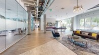 Located at 630 Cotanche St in Greenville, NC, this generously sized single-floor amenity space is thoughtfully designed to blend fitness, work, and leisure into one cohesive environment. The fitness area is a standout feature, partially enclosed in a sleek glass-walled structure that showcases a robust selection of cardio equipment — including treadmills, ellipticals, and stationary bikes — alongside strength training machines, free weights, and benches. Exposed ductwork and soaring ceilings lend an industrial-modern aesthetic throughout, while large floor-to-ceiling windows flood the space with natural light and offer views of the surrounding urban streetscape. An adjacent open exercise zone features full-length mirrors and hardwood-style flooring, providing ample room for functional training. The entertainment lounge seamlessly connects to the fitness area and includes a billiard table, ping pong table, a wall-mounted TV, and comfortable seating arrangements anchored by statement pendant lighting. A vibrant pop-art mural adds bold visual character to the fitness zone. The co-working and social areas feature glass-partitioned meeting rooms, round dining tables with bold yellow accent chairs, and inviting lounge seating — all bathed in natural light, creating a dynamic and energizing atmosphere ideal for both productivity and relaxation.