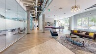 Located at 630 Cotanche St in Greenville, NC, this generously sized single-floor amenity space is thoughtfully designed to blend fitness, work, and leisure into one cohesive environment. The fitness area is a standout feature, partially enclosed in a sleek glass-walled structure that showcases a robust selection of cardio equipment — including treadmills, ellipticals, and stationary bikes — alongside strength training machines, free weights, and benches. Exposed ductwork and soaring ceilings lend an industrial-modern aesthetic throughout, while large floor-to-ceiling windows flood the space with natural light and offer views of the surrounding urban streetscape. An adjacent open exercise zone features full-length mirrors and hardwood-style flooring, providing ample room for functional training. The entertainment lounge seamlessly connects to the fitness area and includes a billiard table, ping pong table, a wall-mounted TV, and comfortable seating arrangements anchored by statement pendant lighting. A vibrant pop-art mural adds bold visual character to the fitness zone. The co-working and social areas feature glass-partitioned meeting rooms, round dining tables with bold yellow accent chairs, and inviting lounge seating — all bathed in natural light, creating a dynamic and energizing atmosphere ideal for both productivity and relaxation.