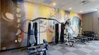 Located at 1179 E Main St in Columbus, Ohio, this fitness center offers a thoughtfully designed workout space within a compact yet efficiently utilized single-floor layout. The room is finished with a two-tone modular carpet tile floor in shades of gray, providing a clean and functional surface throughout. A drop ceiling with recessed lighting and a ceiling fan ensures the space remains well-lit and comfortable during workouts.

One of the most striking design features is a large, vivid mural spanning an entire wall, depicting a colorful, urban-inspired Columbus-themed collage with bold typography and abstract imagery in yellows, blues, and earth tones — giving the space a dynamic and energetic character.

Large windows with horizontal slatted blinds line one wall, allowing natural light to filter in while maintaining privacy, with glimpses of outdoor greenery visible above the blinds. The cardio zone features a row of Spirit treadmills alongside elliptical trainers and a stationary bike. A multi-station cable weight machine anchors the strength training area, complemented by adjustable workout benches and a rack of dumbbells and kettlebells. Additional accessories such as stability balls and resistance equipment are also present. A wall-mounted television and glass entry doors complete this practical, modern fitness facility.