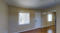 This single-floor space offers a practical and efficient layout designed to maximize usability. The main living area is spacious relative to the overall footprint, featuring warm-toned flooring with a subtle texture that adds character to the room. A large window fitted with vertical blinds allows natural light to filter in, brightening the interior and creating an inviting atmosphere. The front entry door is finished in white with an arched panel detail, providing a clean and classic aesthetic.

The kitchen occupies the right-hand portion of the floor plan and is equipped with a stove, sink, and counter space arranged for convenient use. It transitions smoothly from the living area, making the overall flow of the space feel open and connected. Adjacent to the kitchen is a compact bathroom area, completing the functional zones of the unit.

Warm beige walls are consistent throughout, complementing the wood-toned flooring and contributing to a cohesive, neutral color palette. White baseboards and trim provide crisp contrast against the walls. The layout is thoughtfully organized, with each area clearly defined while maintaining an open feel. Overall, this space offers a straightforward and comfortable environment suited for everyday living.
