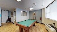 This single-floor recreational facility is thoughtfully designed to offer a variety of leisure and social spaces. Light hardwood flooring runs continuously throughout, creating a warm, cohesive feel across all areas. The layout is open and well-connected, allowing easy flow between zones.

The centerpiece of the space is a dedicated billiards room, featuring a full-size pool table with a green felt surface, a wall-mounted cue rack, and flush panel lighting overhead. Windows with horizontal blinds allow natural light to filter in, while a textured ceiling and blue-gray accent walls add character.

Adjacent to the billiards area is a lounge and waiting zone furnished with burgundy upholstered seating. Sports-themed décor, including a prominent Philadelphia Eagles banner, lends a casual, community-oriented atmosphere. A restroom is conveniently accessible from this corridor, with signage clearly marking its location.

A quieter nook features a dedicated chess area with a wooden chess set displayed on a small table, flanked by two chairs. The walls here are finished with white tile wainscoting below blue-gray paint, and a decorative chess piece wall mural adds a playful touch.

Open common areas with movable partitions and additional seating provide flexible space for gatherings or group activities. The facility is well-lit with recessed panel lighting throughout and includes safety features such as fire extinguishers and alarm systems.