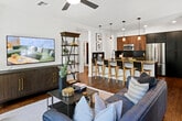 Located at 1901 E Dyer Rd in Santa Ana, California, this single-floor unit blends modern industrial design with warm, eclectic touches. The open-concept kitchen is a standout, equipped with stainless steel appliances including a gas range, built-in microwave, and French door refrigerator, all set against dark cabinetry and navy blue subway tile backsplash. A generous island with a dark countertop and undermount sink is illuminated by a row of Edison-style pendant lights, offering casual seating and a natural flow into the dining area. The dining space features an exposed brick accent wall, large windows that invite abundant natural light, and an eye-catching multi-globe chandelier. Rich hardwood flooring runs throughout, anchoring the cohesive design. The living room continues the industrial-chic aesthetic with another exposed brick wall, a ceiling fan, large windows with outdoor greenery views, and comfortable seating. The bedroom is a serene retreat with a textured accent wall, woven pendant light, and a bright window overlooking the exterior. The bathroom is elegantly appointed with large hexagonal floor tile, a walk-in glass-enclosed shower with white tile surround, black matte fixtures, and built-in shelving. This space masterfully combines raw architectural elements with refined modern finishes.