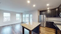 This thoughtfully designed single-floor residence offers a clean, modern aesthetic complemented by quality finishes throughout. The open-concept kitchen and living area serves as the heart of the home, anchored by a large center island topped with speckled granite countertops. Dark espresso cabinetry lines the kitchen walls, paired with stainless steel appliances including a refrigerator, range, and built-in microwave. Rich hardwood flooring flows seamlessly through the main living space, which is bathed in natural light from large windows and a glass door that opens directly onto a private balcony with outdoor views of the surrounding landscape. The bedrooms feature plush carpeting and multiple windows that frame serene, elevated views of greenery and the surrounding area. A hallway connects the rooms, finished with the same warm hardwood flooring and neutral-toned walls. The bathroom showcases a classic black-and-white mosaic tile floor, dark vanity cabinetry with a white countertop, a large mirror, and a full tub-shower combination with white subway tile surround. Throughout the space, recessed lighting, clean white trim, and a neutral color palette create a bright, cohesive, and move-in-ready environment.