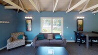 Located at 6921 Salge Dr in Fort Wayne, Indiana, this single-story community clubhouse offers a well-appointed and versatile gathering space designed for both relaxation and productivity. The main gathering area is a standout, featuring soaring vaulted ceilings with exposed white-painted wood beams and a dramatic white brick fireplace as its centerpiece. Bold accent walls in teal and chartreuse green create a vibrant, modern atmosphere throughout the space. A comfortable lounge area with a gray sofa, accent chairs, and a coffee table is bathed in natural light from large windows overlooking the surrounding greenery. Adjacent to the lounge, a dining area with a dark wood table and upholstered chairs provides an ideal setting for meetings or social gatherings. Double glass-paned doors open to an outdoor deck, further enhancing the connection to the outdoors. The clubhouse also includes a fully equipped kitchenette with stainless steel appliances, dark wood cabinetry, and tile flooring. Two dedicated office or workstation areas offer functional spaces for business use, each featuring carpeted flooring, built-in desks, and garden views. The cohesive color palette, decorative pendant lighting, and curated artwork tie the entire space together into an inviting and functional community hub.