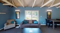 Located at 6921 Salge Dr in Fort Wayne, Indiana, this single-story community clubhouse offers a well-appointed and versatile gathering space designed for both relaxation and productivity. The main gathering area is a standout, featuring soaring vaulted ceilings with exposed white-painted wood beams and a dramatic white brick fireplace as its centerpiece. Bold accent walls in teal and chartreuse green create a vibrant, modern atmosphere throughout the space. A comfortable lounge area with a gray sofa, accent chairs, and a coffee table is bathed in natural light from large windows overlooking the surrounding greenery. Adjacent to the lounge, a dining area with a dark wood table and upholstered chairs provides an ideal setting for meetings or social gatherings. Double glass-paned doors open to an outdoor deck, further enhancing the connection to the outdoors. The clubhouse also includes a fully equipped kitchenette with stainless steel appliances, dark wood cabinetry, and tile flooring. Two dedicated office or workstation areas offer functional spaces for business use, each featuring carpeted flooring, built-in desks, and garden views. The cohesive color palette, decorative pendant lighting, and curated artwork tie the entire space together into an inviting and functional community hub.