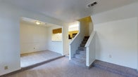 This two-story home offers a straightforward and functional layout designed for comfortable everyday living. The main level centers around a spacious carpeted living area that provides a warm and inviting atmosphere. A centrally positioned staircase with a clean white-painted surround leads to the upper floor, serving as a natural dividing point between the living and kitchen spaces. Adjacent to the living area, a pass-through window opens to the kitchen, which features warm wood-toned cabinetry and bright overhead lighting that enhances visibility throughout the cooking space. The kitchen's layout appears well-organized, with cabinetry lining the walls and ample room for meal preparation. Neutral wall tones throughout the main level create a clean, versatile backdrop suited to a variety of interior styles. The ceiling incorporates a textured finish in select areas, adding a subtle design element to the space. HVAC venting is integrated into the ceiling, ensuring climate comfort across the home. The upper floor, accessible via the carpeted staircase, expands the overall living capacity of the home. Together, both levels form a cohesive and practical residence well-suited for those seeking a multi-level living arrangement.