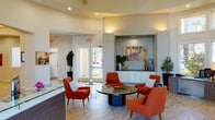 This single-floor leasing office and amenity space is designed with a polished, contemporary aesthetic that creates a warm and professional first impression. The entry foyer opens into a soaring rotunda-style lobby with elevated ceilings, recessed lighting, and an abundance of natural light streaming through large windows and glass double doors that offer views of the surrounding landscaping.

The central lounge area serves as the heart of the space, furnished with bold burnt-orange mid-century modern chairs arranged around a sleek circular table. A built-in display niche with white cabinetry and colorful abstract artwork adds a curated touch to the area. Large-format tile flooring in a diagonal herringbone pattern runs throughout, lending an elegant continuity to the space.

Adjacent to the lobby is the reception and leasing area, featuring a contemporary workstation desk set against a backdrop of floor-to-ceiling windows that flood the space with natural light. A secondary lounge room offers round dining tables with plush velvet seating beneath a statement pendant chandelier, complemented by a built-in TV niche and decorative cabinetry.

A dedicated business center provides a quiet workspace with a built-in desk, multiple computer stations, and soft carpet underfoot. A convenient refreshment station with cabinetry, a coffee maker, and a water cooler rounds out the amenities, ensuring a complete and comfortable visitor experience throughout.