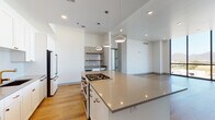 This contemporary single-floor residence is designed with a clean, modern aesthetic highlighted by warm wood-tone flooring, crisp white walls, and high ceilings throughout. The open-concept kitchen serves as the centerpiece of the living space, anchored by a large island with a neutral-toned quartz countertop and an integrated gas cooktop. White shaker cabinetry with gold-toned hardware extends along the perimeter walls, complemented by a chevron-patterned tile backsplash, stainless steel appliances including a built-in microwave, full-size refrigerator, and a wine cooler. Pendant lighting and a decorative patterned tile accent wall with open shelving add character to the space. The living area flows seamlessly from the kitchen and is bathed in natural light from expansive floor-to-ceiling windows, which frame sweeping views of the surrounding cityscape and distant mountains. Sliding glass doors open to a private balcony, further connecting the interior to the outdoors. The bathrooms are finished consistently with gray quartz countertops, white cabinetry with copper-toned hardware, undermount sinks, and backlit mirrors. Additional rooms offer versatile, well-proportioned spaces with ample natural light.
