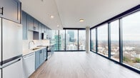 Located in the heart of Midtown Atlanta along the iconic Peachtree Street corridor, this high-rise residence offers a sleek, contemporary living experience defined by soaring floor-to-ceiling windows that frame breathtaking panoramic views of the Atlanta skyline and surrounding cityscape. Light-toned hardwood-style flooring flows throughout, complementing the clean, modern aesthetic.

The open-concept kitchen and living area serves as the heart of the unit, where custom slate-blue shaker cabinetry pairs with white subway tile backsplash, bright quartz countertops, and a full suite of stainless steel appliances. The kitchen opens seamlessly into the expansive living area, which is bathed in natural light and anchored by dramatic city views through the full-height glazed window wall.

The bedroom features its own floor-to-ceiling window with city views, a ceiling fan, and hardwood-style flooring, with access to well-appointed closets offering built-in shelving and hanging rods. The hallway connects the private sleeping quarters to the main living spaces with a clean, minimalist design.

Two bathrooms serve the residence — one with a deep soaking tub and white subway tile surround, and another with a glass-enclosed walk-in shower finished in matching tile. Both feature large-format tile flooring and light wood vanities.