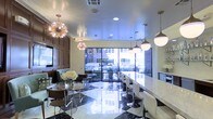 This single-floor space combines bold design elements with functional versatility, creating an inviting environment suited for gatherings or events. The most striking feature is the high-gloss black-and-white checkered marble flooring, which anchors the room with dramatic visual impact and reflects light throughout the interior.

Two distinct seating areas define the layout. A casual lounge zone on one side features a round glass-top table surrounded by modern wire-frame and upholstered chairs, accented by a floral centerpiece. On the opposite side, a long white countertop bar runs the length of the wall, lined with sleek white and chrome bar stools, ideal for dining or socializing. A built-in counter or bar area is partially visible at the far end.

Overhead, a curated mix of pendant lights — including two bold Sputnik-style brass fixtures and several globe and cone pendants — creates a layered, mid-century modern aesthetic. Large floor-to-ceiling windows along the back wall invite abundant natural light and offer views of the surrounding streetscape. The combination of warm tones, reflective surfaces, and eclectic lighting gives the space a polished yet welcoming character.