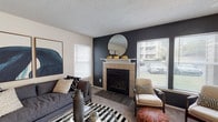 This single-floor residence offers a well-organized layout with distinct living, dining, kitchen, and bedroom spaces. The living room serves as the focal point of the home, showcasing a tile-surround fireplace set against a bold dark accent wall. A round mirror adorns the mantel, while multiple windows with horizontal blinds allow natural light to fill the carpeted space, creating a bright and contemporary atmosphere.

The kitchen is equipped with warm honey-toned wood cabinetry, light stone-look countertops, and white appliances including an electric range and refrigerator. A peninsula with a breakfast bar extending into the adjacent living area provides additional seating and workspace, with a sliding glass door nearby offering access to an outdoor balcony.

The bedrooms feature plush carpeting, ceiling fans, and generous closet space. One bedroom includes a sliding glass door with outdoor views, while another features a striking dark teal accent wall paired with bold graphic artwork, lending a stylish, design-forward character.

The bathroom includes a single-sink vanity with wood cabinetry, a large mirror, and a shower-tub combination. Textured ceilings are consistent throughout the home, contributing to its classic residential character.
