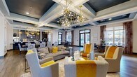 Located at 3200 Brook Rd in Richmond, Virginia, this elegantly appointed single-floor clubhouse offers a generously sized communal space designed to impress. The entry reception area sets the tone with a striking herringbone-patterned wood-look floor, a sculptural front desk, sputnik-style gold pendant lighting, and glass-walled private offices lined with navy blue seating. The grand lounge and community room features dramatic coffered ceilings painted in deep navy and white, adorned with elaborate gold geometric chandelier fixtures that serve as true focal points. Plush seating in warm gold and neutral tones is arranged throughout, while large windows flood the space with natural light and provide views to the outdoor area. A formal dining zone accommodates a large table with patterned upholstered chairs beneath a rectangular chandelier. The full catering kitchen is outfitted with dark cabinetry, stainless steel appliances, quartz countertops, a double sink, and a textured tile accent wall with a mounted television. A dedicated workspace corridor features a live-edge wood built-in counter and large-scale contemporary artwork. Wood-look tile flooring runs consistently throughout, unifying each distinct area of this refined, community-focused facility.