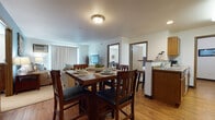 This single-floor unit offers a well-organized, open-concept layout that connects the main living areas seamlessly. The dining area is positioned centrally, accommodating a substantial raised dining table with seating for several, and flows naturally into the adjacent living room. The living room features a calming blue accent wall, vertical blind-covered sliding access, and a wall-mounted air conditioning unit, creating a cozy yet functional retreat.

The kitchen is efficiently designed with warm oak cabinetry providing generous storage, stone-look countertops, and a full suite of white appliances including a range, over-the-range microwave, and refrigerator. A peninsula with bar-height seating separates the kitchen from the dining space, enhancing the open feel. Light hardwood-style flooring runs throughout the main living areas, adding warmth and continuity.

The bedroom is carpeted and comfortably scaled, receiving natural light through a window with horizontal blinds. A flexible secondary room with carpet can serve as a home office or additional bedroom. Two full bathrooms are finished with stone-tile flooring, oak vanities, and light countertops—one featuring a tub-shower combination and the other a spacious vanity with a large mirror.