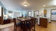 This single-floor unit offers a well-organized, open-concept layout that connects the main living areas seamlessly. The dining area is positioned centrally, accommodating a substantial raised dining table with seating for several, and flows naturally into the adjacent living room. The living room features a calming blue accent wall, vertical blind-covered sliding access, and a wall-mounted air conditioning unit, creating a cozy yet functional retreat.

The kitchen is efficiently designed with warm oak cabinetry providing generous storage, stone-look countertops, and a full suite of white appliances including a range, over-the-range microwave, and refrigerator. A peninsula with bar-height seating separates the kitchen from the dining space, enhancing the open feel. Light hardwood-style flooring runs throughout the main living areas, adding warmth and continuity.

The bedroom is carpeted and comfortably scaled, receiving natural light through a window with horizontal blinds. A flexible secondary room with carpet can serve as a home office or additional bedroom. Two full bathrooms are finished with stone-tile flooring, oak vanities, and light countertops—one featuring a tub-shower combination and the other a spacious vanity with a large mirror.