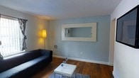 This single-floor unit is organized into distinct functional zones that work together to create a cohesive and practical living environment. The layout, as seen from the floor plan, divides the space into a bedroom on one side and an open living area on the other, with a kitchen and bathroom completing the arrangement.

The living area is finished with rich, dark hardwood flooring that extends throughout much of the unit, lending warmth and a sense of continuity to the space. Walls are painted in a soft blue-gray tone, contributing to a calm and understated atmosphere. A large rectangular mirror with a white frame is mounted prominently on the main wall, reflecting light and enhancing the sense of openness. The room accommodates a seating area with a dark sofa, a coffee table, and a floor lamp that provides warm ambient lighting.

The bedroom features carpeted flooring, offering a softer contrast to the hardwood in the adjacent rooms. The kitchen is compact yet functional, positioned to the rear of the unit. A bathroom completes the layout. Throughout the unit, clean white walls and simple finishes maintain a neutral, easy-to-customize aesthetic.