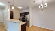 This single-floor space showcases a thoughtfully designed open-concept layout that seamlessly connects the kitchen and dining areas. The kitchen is equipped with a full suite of stainless steel appliances, including a French-door refrigerator, electric range, and over-the-range microwave. Rich, dark espresso cabinetry provides ample storage and creates a striking contrast against the light granite-style countertops. A breakfast bar with a light blue accent panel extends from the counter, offering casual seating and additional workspace. Light hardwood-style flooring runs throughout, enhancing the bright and airy atmosphere of the space. Overhead, a combination of pendant lighting above the kitchen area and a decorative chandelier near the dining space adds both functional illumination and stylish detail. The neutral white walls provide a clean, versatile backdrop, while standard ceiling height keeps the space feeling comfortable and well-proportioned. The open flow between the kitchen and adjacent dining area makes the layout practical for everyday living and entertaining.