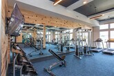 Located at 5030 W Broad St in Sugar Hill, GA, this well-appointed fitness center delivers a comprehensive workout facility designed with both function and aesthetics in mind. The single-floor space is thoughtfully laid out to accommodate a wide range of exercise needs, from cardiovascular training to strength conditioning.

The facility's architectural character is defined by its striking coffered ceilings finished in deep navy blue, which contrast elegantly with the warm, light-toned accent wall featuring a dynamic mosaic-style pattern in muted reds, blues, and naturals — a nod to the Sugar Hill community identity. Structural columns with wood accents add a refined, modern-industrial feel throughout the space.

The cardio zone is generously equipped with treadmills, ellipticals, stair climbers, and a rowing machine, all positioned along a wall of large windows that flood the area with natural light and offer open views of the surrounding landscape. The strength training section features a robust selection of cable machines, multi-station weight systems, free weights, kettlebells, medicine balls, and battle ropes, catering to varied training styles.

Soft blue-toned carpet tiles cover the floor throughout, and recessed and pendant lighting ensure the space is well-lit at all times. A built-in cabinetry unit with a countertop provides a convenient amenity station near the entrance, completing this polished and fully equipped fitness destination.