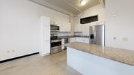 This two-bedroom, two-bathroom unit at 736 Federal Street in Davenport, Iowa offers a distinctive industrial-modern aesthetic across a single floor. The open-concept kitchen serves as a centerpiece, featuring white shaker cabinetry, granite countertops, a stainless steel appliance suite including a French door refrigerator, electric range with over-the-range microwave, and dishwasher, plus a central island for additional prep and dining space. Polished concrete floors and exposed ductwork run throughout, reinforcing the space's urban loft character. High, narrow black-framed windows allow natural light to filter in while maintaining a clean, modern silhouette on the walls. Both bedrooms are generously proportioned, with the primary bedroom offering a comfortable retreat. Each bathroom is finished with white vanity cabinetry, granite countertops, a medicine cabinet with mirror, and tile flooring. The full bathroom includes a tub-shower combination. Two closets provide ample storage. A connecting hallway ties the layout together efficiently. Bold black door frames and base trim provide striking contrast against the neutral white walls, lending the space a cohesive, contemporary industrial character throughout.