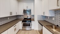 Located at 10125 Palms Blvd in the Palms neighborhood of Los Angeles, this single-story residence offers a well-appointed and thoughtfully designed living space. The kitchen is a standout feature, showcasing white shaker-style cabinetry with brushed nickel hardware, dark countertops, a subway tile backsplash, and stainless steel appliances including a gas range, over-the-range microwave, and dishwasher. Light wood-style flooring runs throughout the main living and kitchen areas, providing a warm and cohesive aesthetic. A well-lit hallway, accented by decorative pendant lighting and a coffered ceiling detail, connects the living spaces and leads toward the bedrooms. The bedrooms are finished with plush wall-to-wall carpeting, crown molding, ceiling fans, and plantation shutters that allow for natural light and privacy control. Neutral, cool-toned paint throughout the bedrooms creates a calm and restful atmosphere. The bathroom features the same light wood flooring as the main areas, with clean white finishes. Crown molding is present in multiple rooms, adding a refined architectural touch. Overall, the space presents a clean, modern interior with quality finishes suited for comfortable everyday living in a prime Los Angeles location.