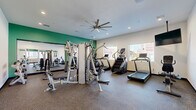 This dedicated fitness center offers a thoughtfully designed exercise space across a single floor. The room is finished with durable rubber flooring suited for high-traffic workout use, white walls accented by a bold green feature wall, and recessed lighting complemented by a large ceiling fan that promotes air circulation during workouts. A full-length mirror spans one wall, visually expanding the space and allowing users to monitor their form. Natural light enters through several windows, creating a bright and energizing atmosphere. The equipment layout is organized to maximize usability, featuring a multi-station cable and strength machine for a comprehensive range of resistance exercises, along with free weights and a dedicated bench. Cardio equipment includes treadmills, a stair climber, and stationary bikes, catering to a variety of fitness routines. A wall-mounted television is also present for entertainment during workouts. Glass-panel entry doors with a view into an adjacent corridor contribute to an open, accessible feel. The overall design balances functionality and comfort, providing users with a well-rounded facility for both cardiovascular and strength training workouts in a clean, organized environment.
