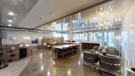 The Club Room is a thoughtfully designed communal space that seamlessly blends elegance with functionality across a single open floor plan. Polished concrete floors reflect the abundant natural light streaming in through floor-to-ceiling windows along the perimeter, creating a bright and dynamic atmosphere throughout the day.

The space is organized into several distinct zones. A central dining area features a large rectangular white-topped table surrounded by dark wood chairs, suitable for group gatherings or collaborative use. Adjacent to this, a built-in bar with sleek counter seating provides a dedicated space for socializing, framed by the expansive window wall beyond.

A lounge area offers plush, curved upholstered seating arrangements ideal for relaxed conversation, while a more intimate built-in banquette with colorful accent pillows anchors a quieter corner alongside curated wall art and decorative paneling.

Overhead, an eclectic mix of designer pendant lights, a sputnik chandelier, and a multi-arm brass fixture add layers of visual interest and warm illumination. The combination of refined finishes, varied seating arrangements, and generous natural light gives the Club Room a welcoming yet upscale character.