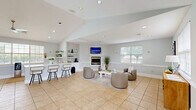 Located at 100 Marshview Dr in Newport News, Virginia, this single-floor community space offers a bright and inviting atmosphere designed for comfort and functionality. The open layout seamlessly connects a living area and dining room, all finished with neutral light gray walls, crisp white wainscoting, and warm beige tile flooring throughout.

The living area is anchored by a clean-lined fireplace with a wall-mounted television above, flanked by plush rounded armchairs arranged around a contemporary white coffee table. Recessed lighting and multiple windows dressed with white horizontal blinds flood the space with natural light and offer pleasant views of the surrounding greenery.

The dining area features a rustic wood table paired with modern gray chairs, positioned beside a large interior window that overlooks an adjacent fitness room — a thoughtful design detail that adds visual connectivity. An exterior glass-panel door provides direct outdoor access, further enhancing the bright, airy feel.

A reception-style zone near the entrance includes a warm wood credenza, comfortable seating, and bold abstract wall art that adds a contemporary touch. Vaulted ceiling sections lend architectural interest and a sense of openness. Together, the cohesive design, abundant natural light, and well-appointed furnishings create a polished, welcoming environment.