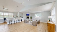 Located at 100 Marshview Dr in Newport News, Virginia, this single-floor community space offers a bright and inviting atmosphere designed for comfort and functionality. The open layout seamlessly connects a living area and dining room, all finished with neutral light gray walls, crisp white wainscoting, and warm beige tile flooring throughout.

The living area is anchored by a clean-lined fireplace with a wall-mounted television above, flanked by plush rounded armchairs arranged around a contemporary white coffee table. Recessed lighting and multiple windows dressed with white horizontal blinds flood the space with natural light and offer pleasant views of the surrounding greenery.

The dining area features a rustic wood table paired with modern gray chairs, positioned beside a large interior window that overlooks an adjacent fitness room — a thoughtful design detail that adds visual connectivity. An exterior glass-panel door provides direct outdoor access, further enhancing the bright, airy feel.

A reception-style zone near the entrance includes a warm wood credenza, comfortable seating, and bold abstract wall art that adds a contemporary touch. Vaulted ceiling sections lend architectural interest and a sense of openness. Together, the cohesive design, abundant natural light, and well-appointed furnishings create a polished, welcoming environment.