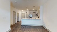 This single-floor space showcases a clean, modern aesthetic defined by an open-concept layout that seamlessly connects the kitchen and living areas. The kitchen is a standout feature, equipped with stainless steel appliances including a French door refrigerator, built-in microwave, and range. White shaker-style cabinetry pairs elegantly with a classic white subway tile backsplash, while a peninsula with a white countertop provides additional workspace and casual seating opportunities. Three brushed metal pendant lights hang above the peninsula, adding a contemporary decorative touch complemented by recessed ceiling lighting throughout. Rich, dark hardwood-style flooring extends across the entire space, creating a warm contrast against the crisp white walls and cabinetry. The open layout allows for a natural flow between the kitchen and the adjacent living area, enhancing the sense of spaciousness. Multiple doors visible from the main living space suggest well-distributed access to additional rooms, while the bright, neutral palette and modern finishes give the space a fresh, move-in-ready appeal.