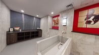 This single-floor space comprises two distinct areas with notably different design aesthetics. The pet grooming wash area is the standout feature, showcasing a contemporary and thoughtfully designed environment. A large, walk-in style bathing tub with an integrated handheld sprayer serves as the focal point, surrounded by elegant stone-look tile cladding the walls. Bold red accent walls provide a striking contrast, complemented by a whimsical tiled mosaic portrait of a Corgi. A wall-mounted light fixture adds functional illumination, while recessed ceiling lights enhance the modern atmosphere. Along one wall, a built-in wooden shelving unit with open cubbies and a countertop work surface provides organized storage. Dark vertical mosaic tile on the opposing wall adds textural depth to the space.

The secondary bathroom presents a more traditional character, with warm cream-toned walls and classic wood cabinetry beneath a white sink vanity. A framed mirror with a distressed wood surround adds a rustic touch above the sink. A toilet with a small wooden rack mounted above completes the functional layout. Brass-finished switch plates and outlet covers carry through a cohesive, traditional hardware aesthetic throughout this area.