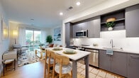 This well-designed one-bedroom, one-bathroom residence at 111 E Broad St in Falls Church, Virginia offers a thoughtfully laid-out single-floor plan. The open-concept kitchen is a standout feature, showcasing sleek charcoal-gray cabinetry extending to the ceiling, white quartz countertops, stainless steel appliances including a refrigerator, range, microwave, and dishwasher, and a central island with bar seating — ideal for casual dining and entertaining. Light wood-tone flooring flows throughout the main living areas, adding warmth to the contemporary design. The living room is bright and inviting, benefiting from large sliding glass doors that open to a private balcony, allowing abundant natural light and outdoor views. The bedroom offers plush carpeting, a generous window with exterior views, and direct access to a dedicated walk-in closet, providing ample storage. Additional closets and a utility room further enhance the functional storage throughout the unit. The bathroom features classic black-and-white basketweave tile flooring, white subway tile surrounding a full soaking tub and shower combination, and a dark-toned vanity. A hallway connects the living spaces cohesively. The overall ambiance is clean, modern, and bright, reflecting a contemporary residential aesthetic suited for comfortable urban living in Falls Church.