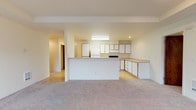 This single-floor residence at 13107 East Mission Avenue in Spokane, Washington offers a practical and well-organized layout with clearly defined living spaces throughout. The living room is generously proportioned, featuring neutral-toned carpet, a wall-mounted air conditioning unit, and a sliding glass door that opens to a private balcony overlooking lush evergreen trees, allowing natural light to fill the space. A tray ceiling detail adds a subtle architectural element to the room.

The kitchen is open and functional, positioned behind a half-wall partition that separates it from the living area. It features light wood laminate flooring, white cabinetry with wood-trimmed accents, ample counter space with neutral laminate countertops, a white refrigerator, and an electric range. The extended counter peninsula provides additional workspace and a visual connection to the main living area.

The bedrooms are carpeted and finished with neutral walls, each equipped with baseboard heating and windows that frame views of the surrounding neighborhood and tree canopy. The bathroom includes a full tub and shower combination with textured gray surround panels, a toilet, and tile flooring. A hallway with wood-toned doors connects the private areas of the unit, completing the straightforward and functional floor plan.