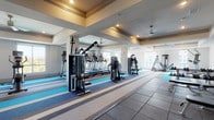 This modern fitness center is thoughtfully designed to accommodate a full range of workout activities across a single, expansive floor. The space is organized into distinct functional zones, each tailored to a specific type of exercise. The cardio area is lined with treadmills, ellipticals, and stationary bikes, all equipped with individual screens and positioned near large windows that invite abundant natural light and outdoor views. The strength training section features a comprehensive selection of cable and weight machines, along with dumbbell racks displayed in front of floor-to-ceiling mirrors that amplify the sense of space. A dedicated free weights area includes adjustable benches and a full dumbbell set for versatile training options. A separate, open stretching and movement area is finished with a distinctive multi-tone striped carpet in blue, grey, and beige tones, providing a clean and energizing aesthetic. A wall-mounted television is positioned in this open zone for entertainment or guided workouts. Throughout the facility, tray ceilings with recessed lighting and ceiling fans maintain comfort and visual sophistication. Structural columns punctuate the layout while supporting the architectural design. Glass-panel doors connect the zones seamlessly, ensuring an open, airy flow across the entire facility.