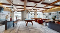 This single-floor amenity lounge is thoughtfully designed to serve as a dynamic social hub, blending upscale aesthetics with recreational functionality. The space is defined by its standout coffered ceiling, accented with rich wood beams and complemented by an eclectic mix of pendant lighting, including cage-style and lantern-inspired fixtures that add warmth and visual interest throughout.

The main gathering area centers around a full-size pool table with a slate-blue felt surface and a nearby shuffleboard table, both positioned to take full advantage of the abundant natural light streaming through oversized floor-to-ceiling windows. These windows frame views of the surrounding exterior and fill the space with daylight, enhancing the open, airy feel.

A comfortable lounge zone features a large dark sectional sofa accented with warm orange and neutral throw pillows, arranged around a wood coffee table against a dramatic exposed brick accent wall with framed artwork. Clusters of tufted burnt-orange swivel chairs provide additional casual seating near the windows.

A semi-private dining nook offers a round wood table with upholstered navy chairs, set against a brick feature wall with curated artwork and decorative sconce lighting. The large-format tile flooring and layered area rugs tie the various zones together seamlessly.
