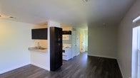 This single-floor space showcases an open-concept design that seamlessly connects the kitchen and living area, creating a cohesive and functional layout. The kitchen is outfitted with dark espresso cabinetry, including a tall pantry-style cabinet that provides generous storage. A granite-patterned countertop with an integrated sink adds a practical yet stylish touch, while white appliances — including an electric range and over-the-range microwave — offer a clean contrast against the darker cabinetry. A half-wall peninsula separates the kitchen from the adjoining living space, maintaining an open feel while defining each zone. Throughout the space, dark hardwood-style flooring runs continuously, lending warmth and visual cohesion. The walls are finished in a soft neutral tone, complemented by crisp white trim and baseboards. Ceiling-mounted HVAC vents suggest a central air system. Toward the rear of the unit, two white paneled doors are visible, likely leading to private rooms or a hallway, hinting at a well-organized floor plan. The overall ambiance is clean, modern, and move-in ready.